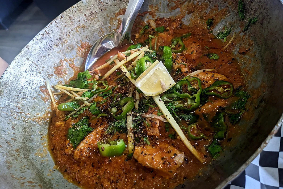 Chicken karahi in a steel karahi pan, topped with green chilies, ginger strips and a lemon wedge