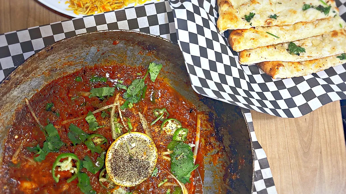 Top-down table spread: a karahi pan of curry with lemon and green chilies, naan in a checkered basket and a plate of biryani