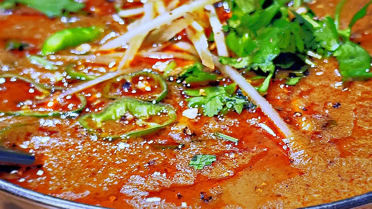 Close-up of a rich curry in a hammered steel bowl, topped with ginger strips, green chilies and fresh coriander